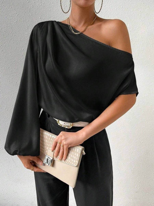 Woman wearing a black one-shoulder top and holding a beige clutch against a neutral background