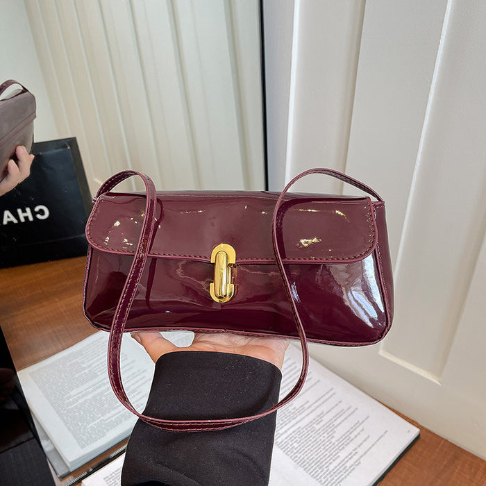 A glossy burgundy patent leather handbag with a gold clasp and matching shoulder strap, held by a hand over an open book in an indoor setting.