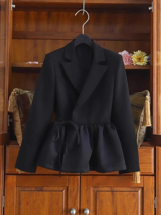 A black tailored blazer-style jacket with a wrap front and drawstring waist, hanging on a hanger in front of a wooden cabinet.