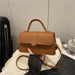 A tan faux-leather handbag with a top handle and shoulder strap, held by a hand over a white fabric surface in an indoor setting.