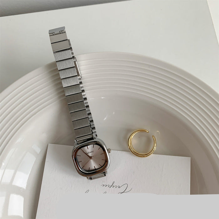 A silver-toned metal link watch with a square analog face and matching metal bracelet, styled on a white plate with a gold ring and a card underneath.