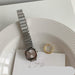 A silver-toned metal link watch with a square analog face and matching metal bracelet, styled on a white plate with a gold ring and a card underneath.
