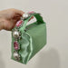 Green satin handbag with decorative handle featuring pearls and pink stones held by a hand against a neutral background.
