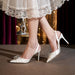 White high-heeled shoes with decorative elements worn with a lace-trimmed dress.