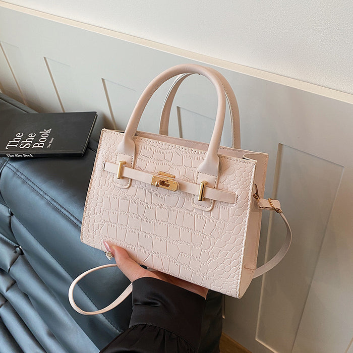 A light pink crocodile-embossed handbag with gold-tone hardware and double top handles, held by a person in a black sleeve against a modern interior background with a dark leather sofa and a book.