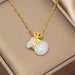 A gold chain necklace with a pendant featuring a carved white jade-like rabbit and a small gold heart-shaped charm with tiny clear stones, displayed inside a beige jewelry box.