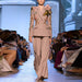Model walking on a fashion runway wearing a beige suit with floral embroidery.