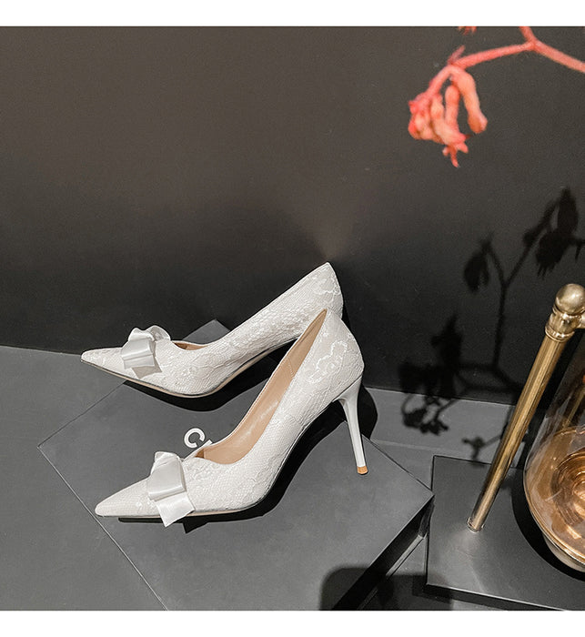 White lace high-heeled shoes with bow details on a dark surface.
