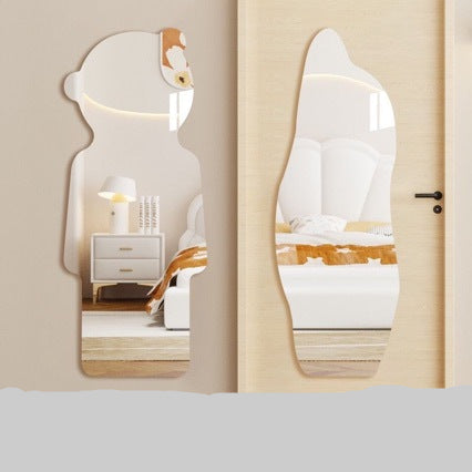 Two irregularly shaped wall mirrors with rounded edges are shown mounted on a beige wall next to a door. The left mirror is shaped like a cartoon character head and reflects a bedroom scene with a white nightstand and lamp.