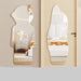 Two irregularly shaped wall mirrors with rounded edges are shown mounted on a beige wall next to a door. The left mirror is shaped like a cartoon character head and reflects a bedroom scene with a white nightstand and lamp.