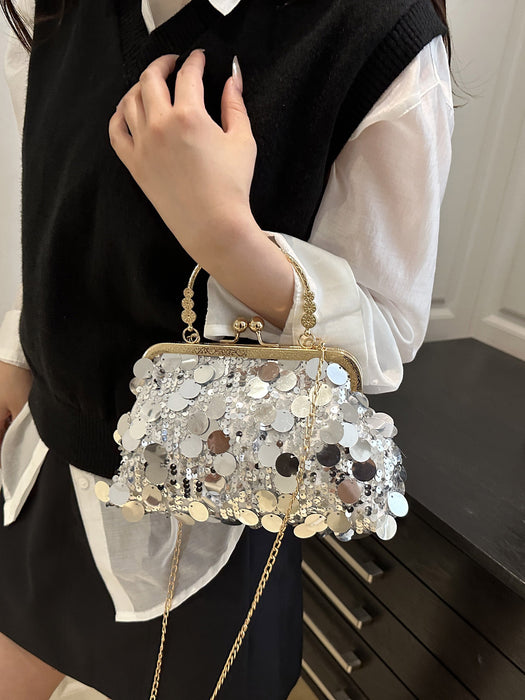 A close-up of a person wearing a black sweater vest over a white blouse, holding a small structured handbag covered in reflective silver and white disc embellishments with a gold-tone frame and chain strap.