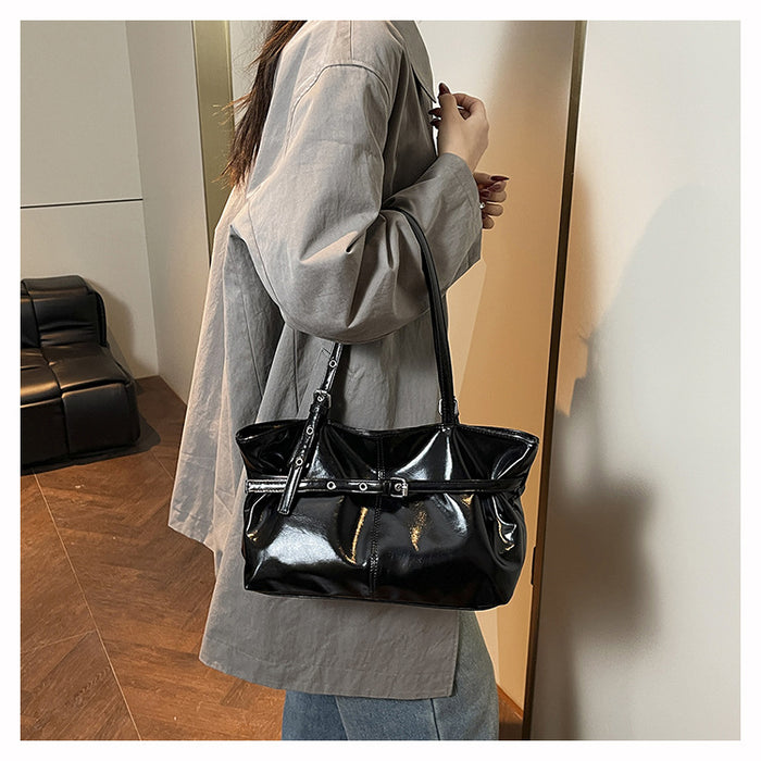 A person wearing an oversized light gray shirt carries a shiny black shoulder bag with a structured top handle and a wide strap detail across the front, photographed indoors near a mirror.