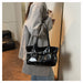 A person wearing an oversized light gray shirt carries a shiny black shoulder bag with a structured top handle and a wide strap detail across the front, photographed indoors near a mirror.