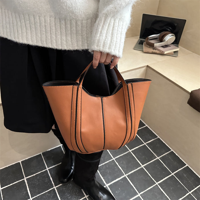 A person wearing a white knit sweater, black skirt, and black knee-high boots holds a tan leather bucket tote bag with black piping details in a modern indoor setting with a black and white tiled floor.