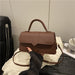 A brown faux-leather handbag with a top handle and shoulder strap, held by a person's foot on a white textured fabric surface.