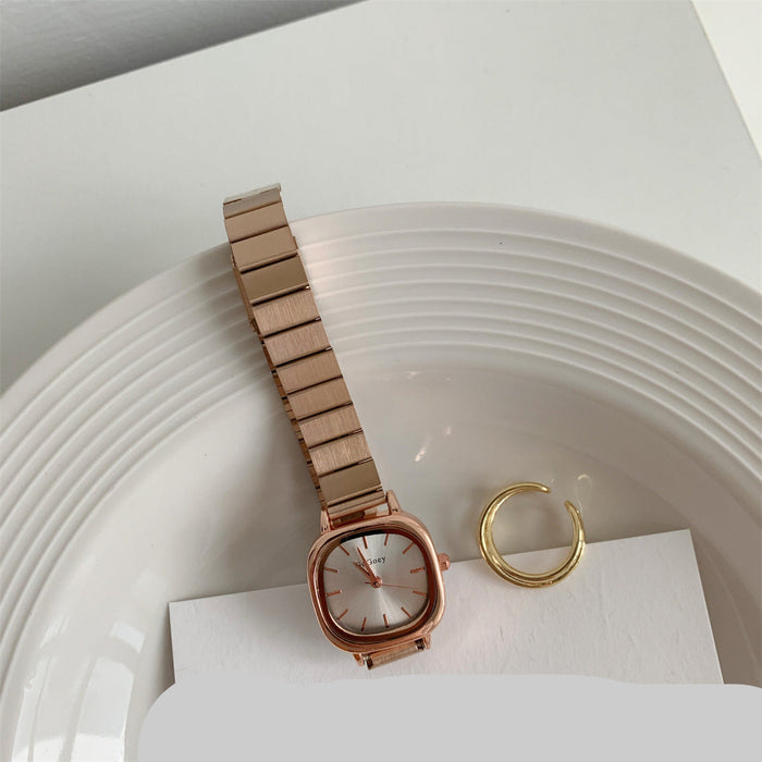 A rose gold-toned women's watch with a square bezel and linked metal bracelet, styled on a white plate with a gold ring and a card.