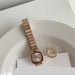 A rose gold-toned women's watch with a square bezel and linked metal bracelet, styled on a white plate with a gold ring and a card.