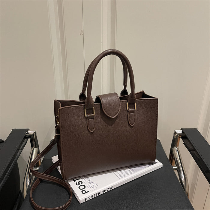 A dark brown textured leather tote bag with double top handles and a detachable shoulder strap, resting on a white book on a black table.
