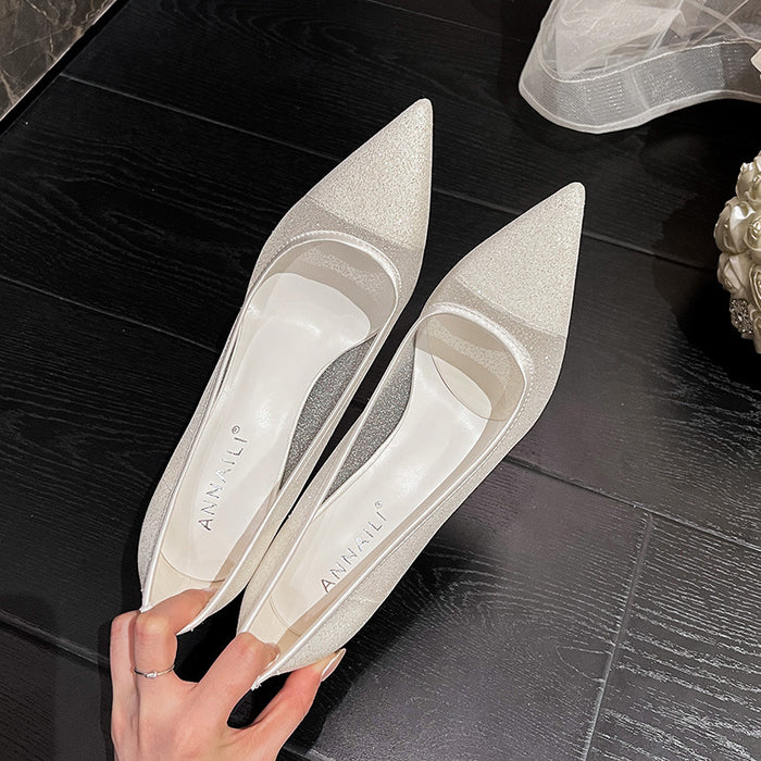 Pair of silver high-heeled shoes held by a hand on a dark wooden floor.