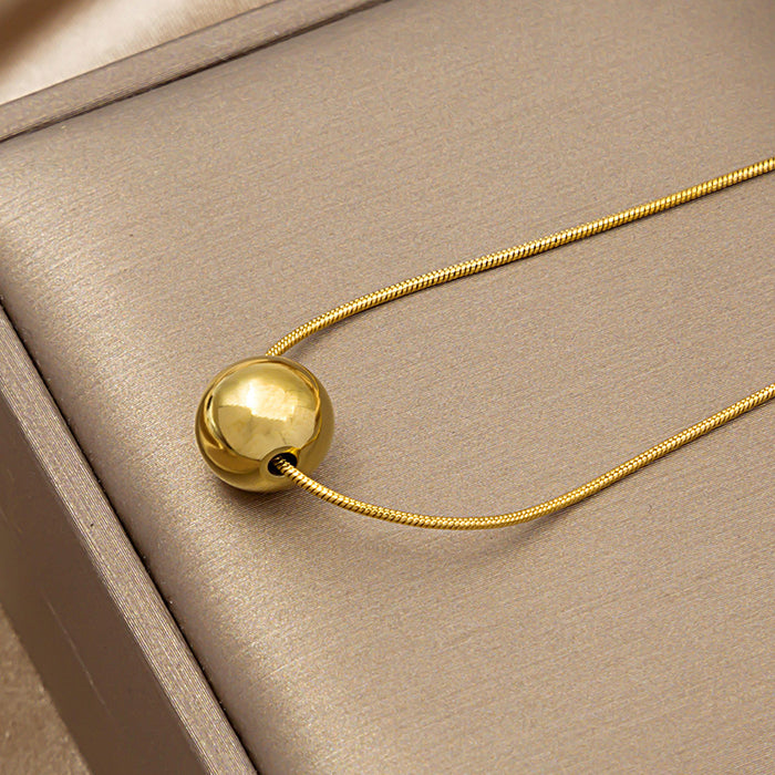 A gold ball pendant necklace on a delicate gold snake chain, displayed on a textured beige jewelry box.