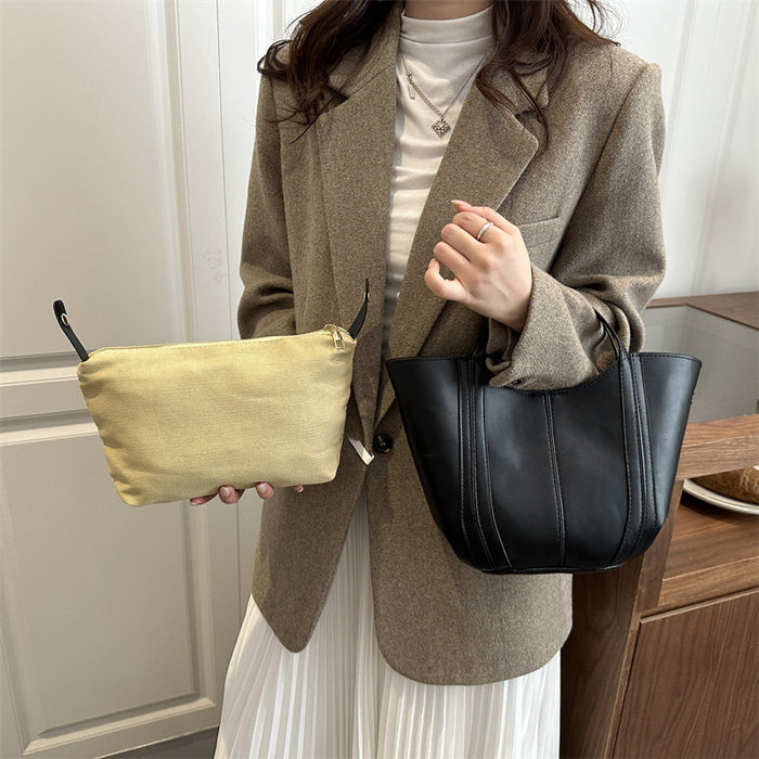 A woman wearing a taupe blazer and white pleated skirt holds a black leather tote bag and a light beige fabric pouch in a home interior setting.