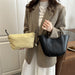 A woman wearing a taupe blazer and white pleated skirt holds a black leather tote bag and a light beige fabric pouch in a home interior setting.