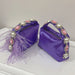 Two purple handbags with decorative handles featuring pearls and feathers on a white surface.