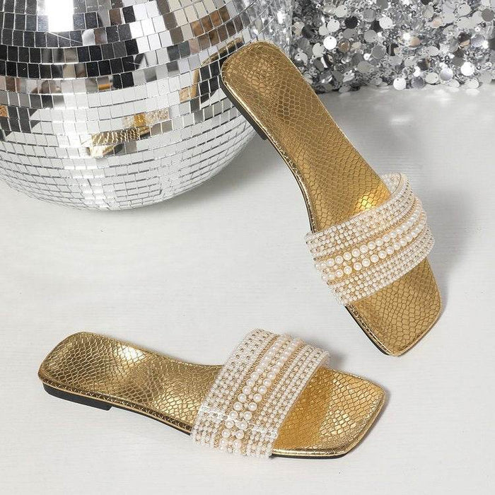 Gold sandals with rhinestone straps next to a disco ball on a reflective surface