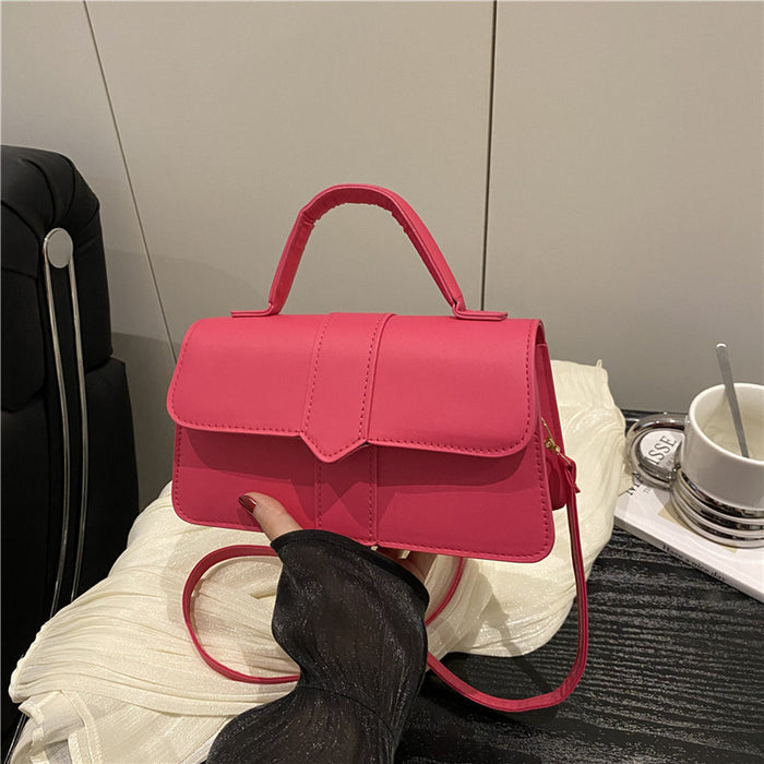A bright pink structured handbag with a top handle and thin shoulder strap, held by a hand over a white textured blanket in an indoor setting.