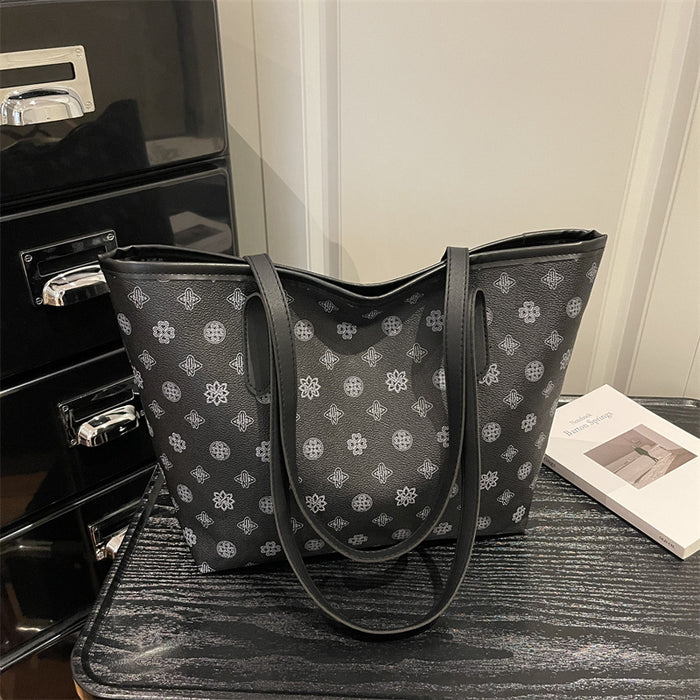 A black tote bag with a repeating white geometric monogram pattern, resting on a dark textured surface next to a black cabinet and a white book.