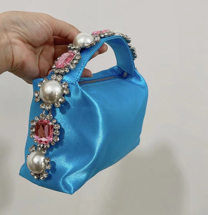 Blue handbag with decorative handle featuring pearls and pink stones held by a hand against a neutral background