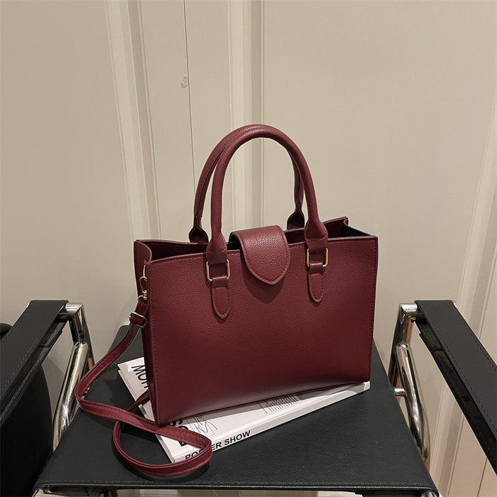 A burgundy pebbled leather tote bag with double top handles and a detachable shoulder strap, resting on a black table with a magazine.