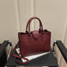 A burgundy pebbled leather tote bag with double top handles and a detachable shoulder strap, resting on a black table with a magazine.