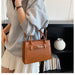 A woman wearing a gray sweatshirt and pleated white skirt carries a brown crocodile-embossed leather handbag with gold-tone hardware in a modern indoor setting.
