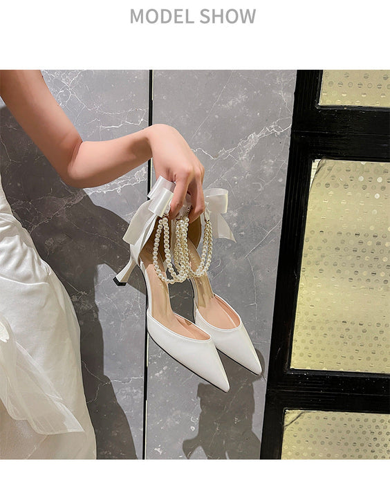 White high-heeled shoes with pearl details held by a person against a marble wall.