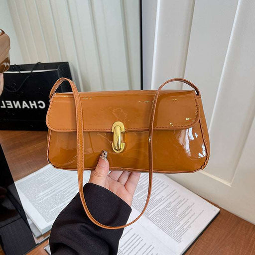 A glossy tan patent leather shoulder bag with a gold clasp, held by a hand over an open book with a Chanel bag in the background.