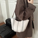 A person wearing a brown coat carries a white tote bag with black vertical stripes and a thin shoulder strap in an indoor setting.