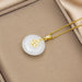 A gold pendant necklace with a round white jade-like disc featuring a gold Chinese character emblem, displayed on a beige jewelry box.
