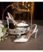 White high-heeled shoes with decorative elements on a decorative stand with flowers in the background.