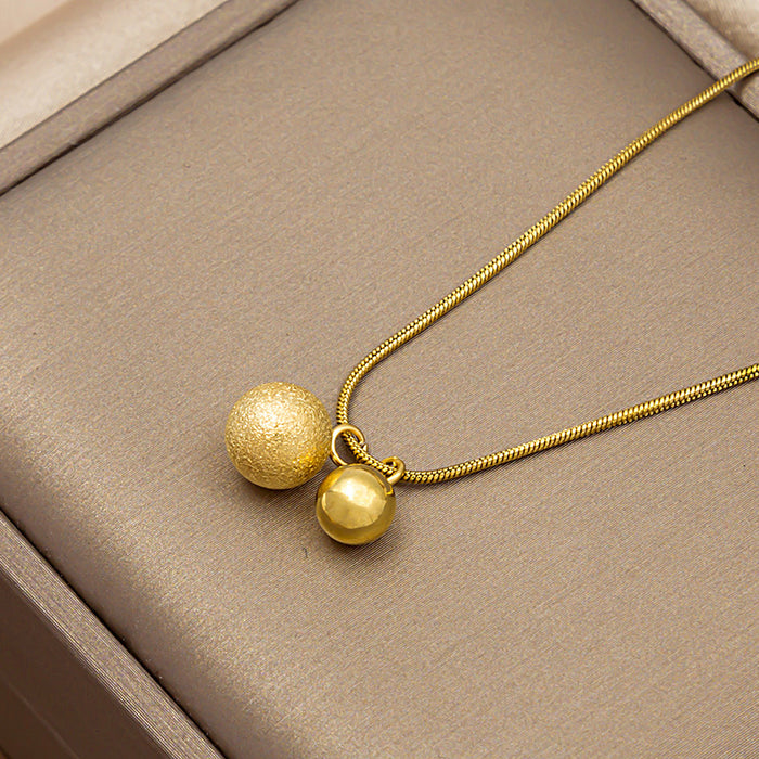 A gold necklace with two spherical pendants, one textured and one smooth, displayed on a beige jewelry box.