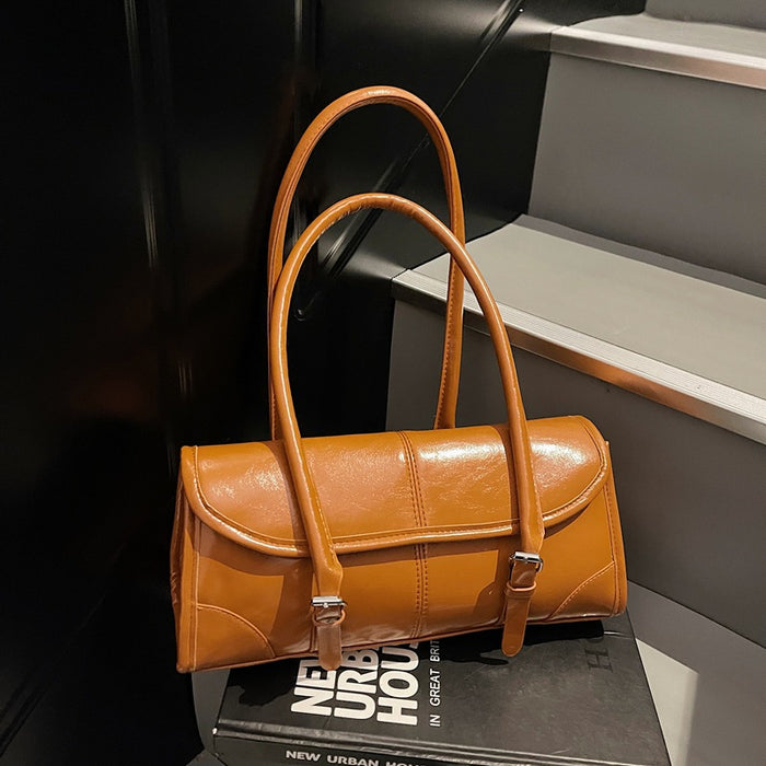 A tan leather handbag with double top handles and silver-tone buckle details, resting on a black book against a dark staircase background.