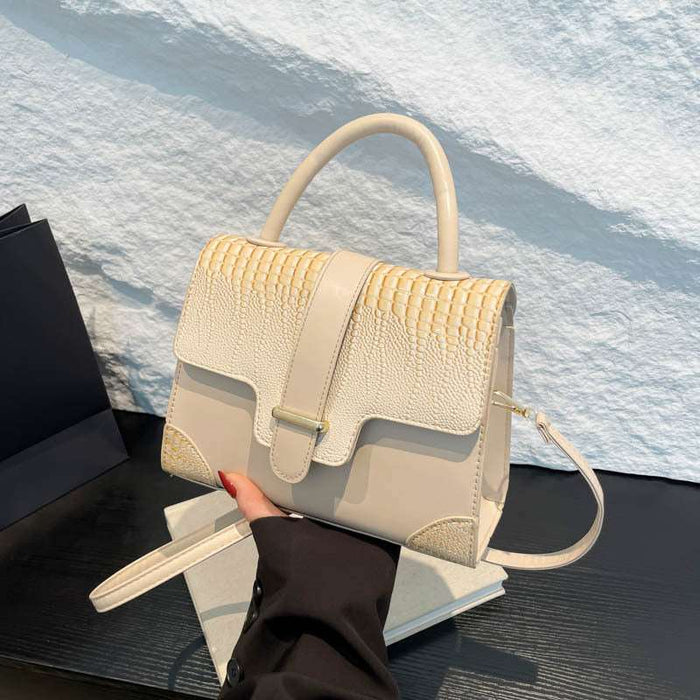 A structured cream-colored handbag with a mix of textured crocodile-embossed and smooth leather panels, featuring a top handle, front flap with a buckle strap, and a detachable shoulder strap. A hand with red nail polish holds the bag, which is resting on a white book against a white textured wall background.