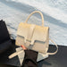 A structured cream-colored handbag with a mix of textured crocodile-embossed and smooth leather panels, featuring a top handle, front flap with a buckle strap, and a detachable shoulder strap. A hand with red nail polish holds the bag, which is resting on a white book against a white textured wall background.