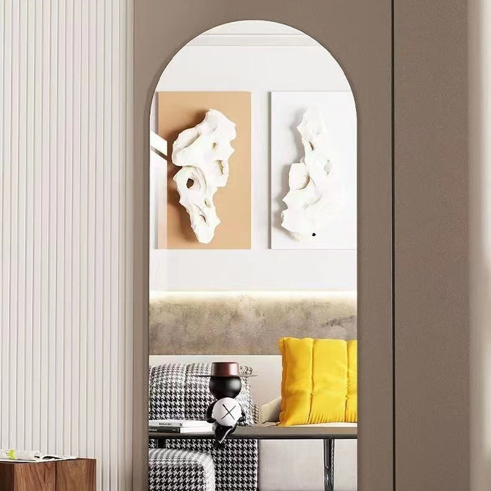 An arched wall mirror with a slim frame hangs on a taupe wall, reflecting a modern living room interior with abstract wall art, a sofa, and a yellow pillow.
