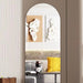 An arched wall mirror with a slim frame hangs on a taupe wall, reflecting a modern living room interior with abstract wall art, a sofa, and a yellow pillow.