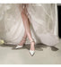 White high-heeled shoes with decorative straps worn by a person in a white dress.