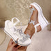 White wedge sandals with silver floral embellishments held by a hand against a neutral background