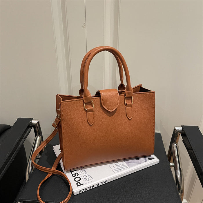 A structured tan leather handbag with double top handles and a shoulder strap, displayed on a black chair in front of a light-colored wall.