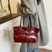 A person wearing an oversized light gray coat holds a shiny deep red tote bag with silver-tone hardware in an indoor room setting.