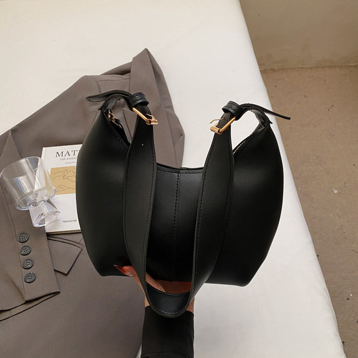A black faux-leather shoulder bag with a structured, slouchy shape and gold-tone hardware, held by a hand in front of a taupe blazer and a clear glass on a white surface.